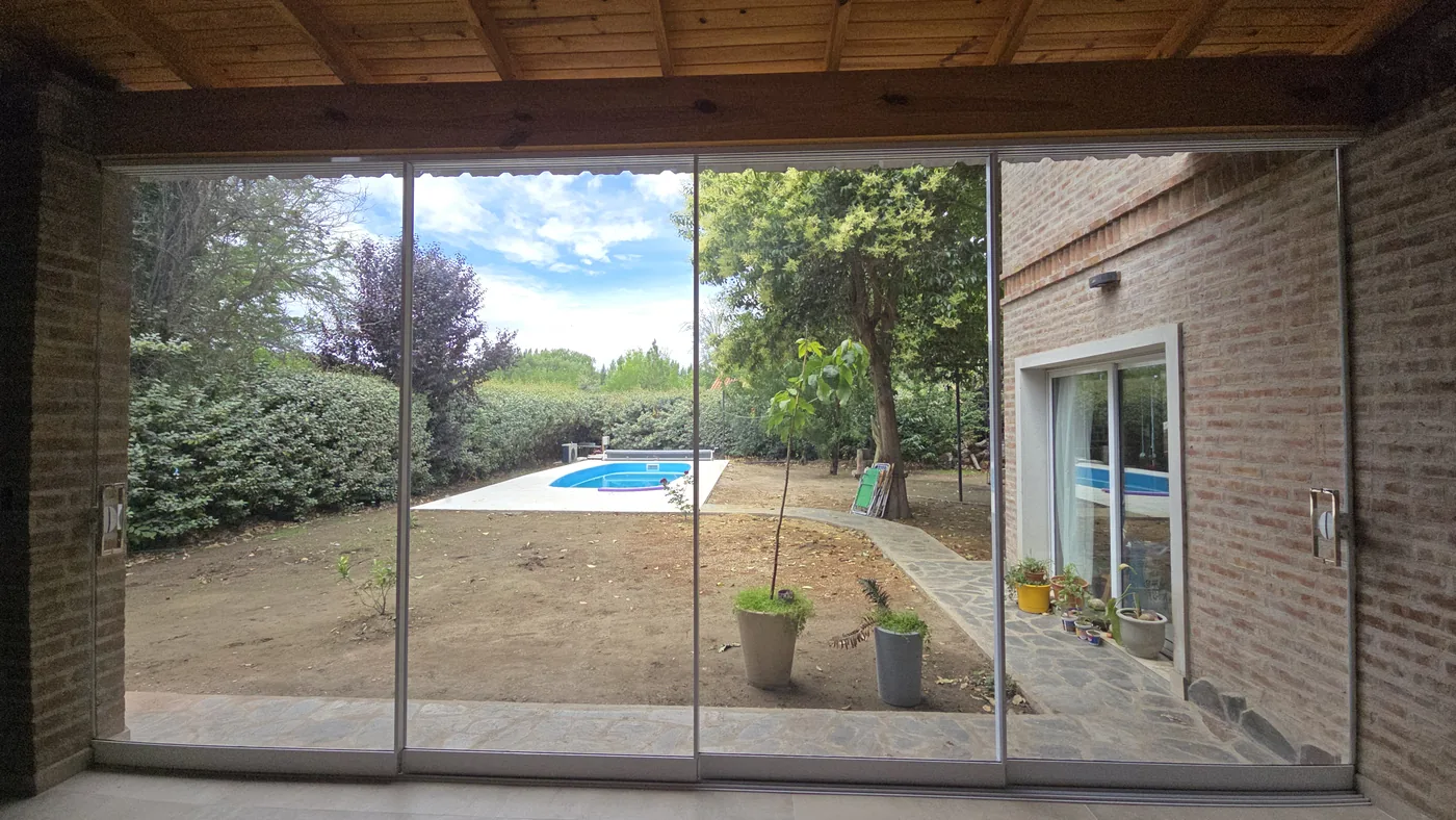 Cerramiento minimalista de vidrio con perfilería de aluminio natural bajo galería de madera, abierto a la pileta - obra de JN Aluminio en Neuquén